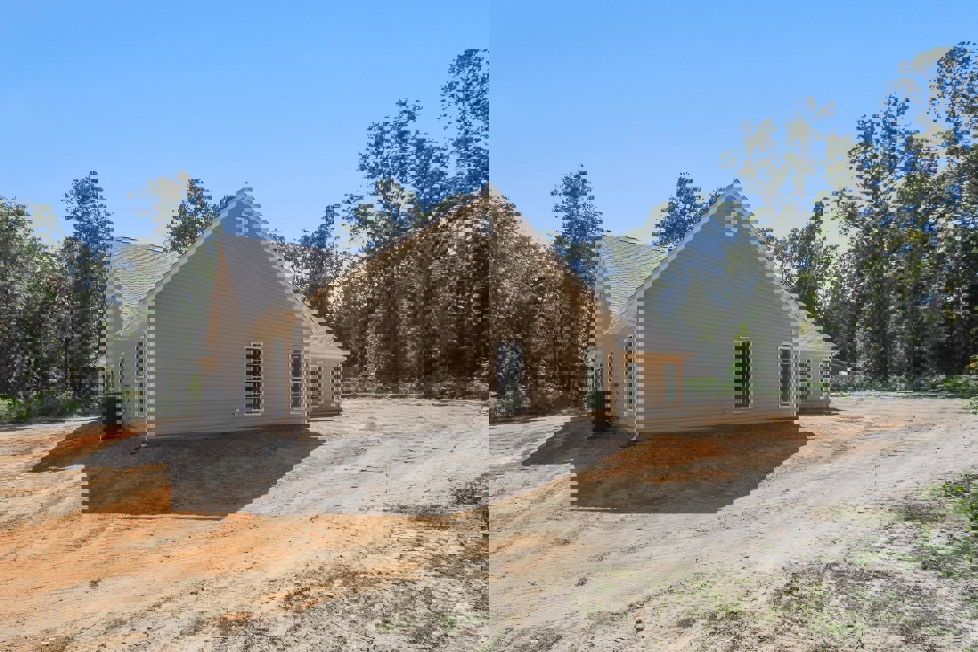 Partially built house with shingled roof, white-framed window, surrounded by dirt lot and scattered trees, black metal utility box with hose in foreground
