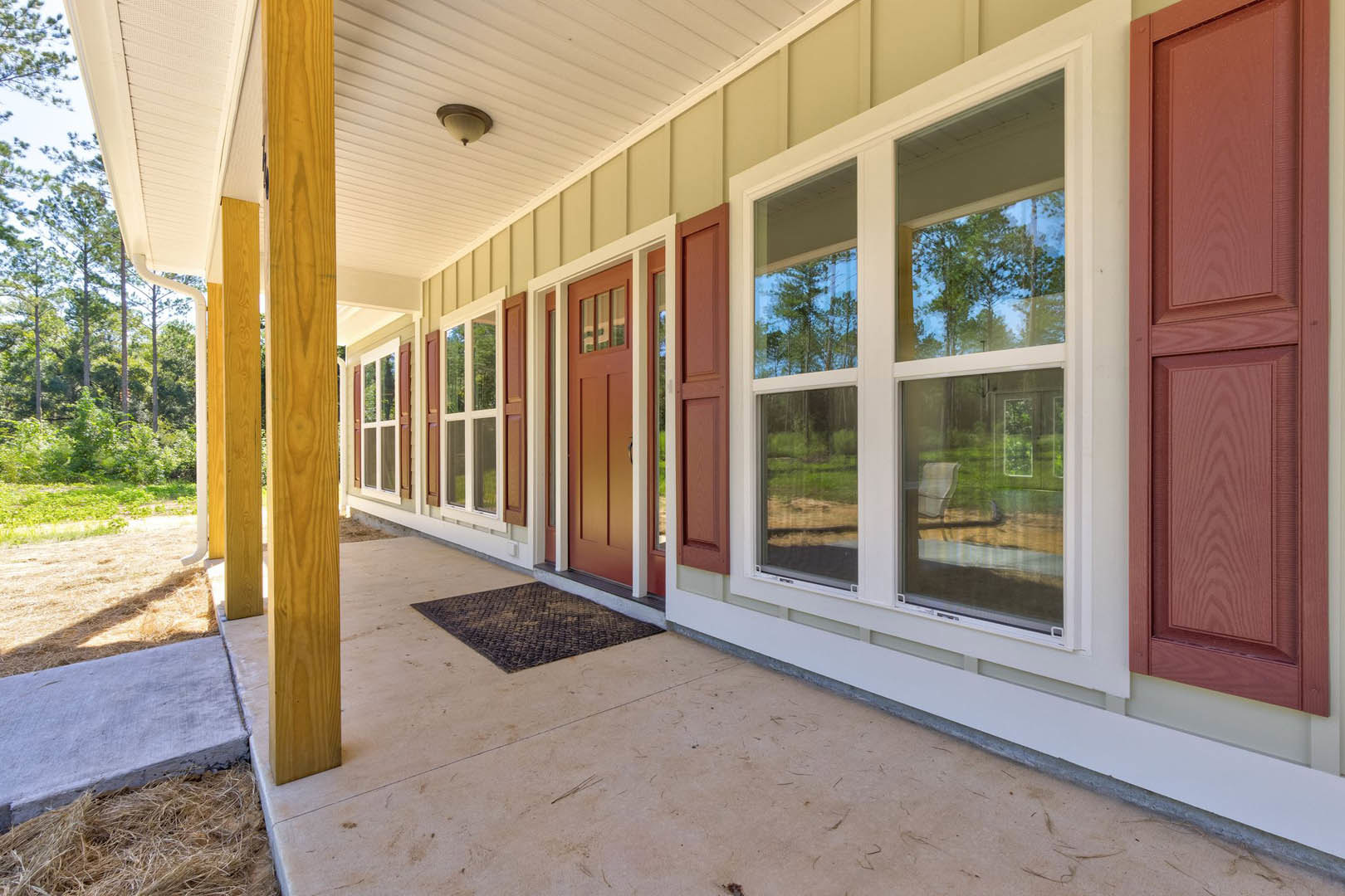 Wide front porch with wood decking, white trim, glass-paneled door, welcome mat, and large window overlooking leafy trees