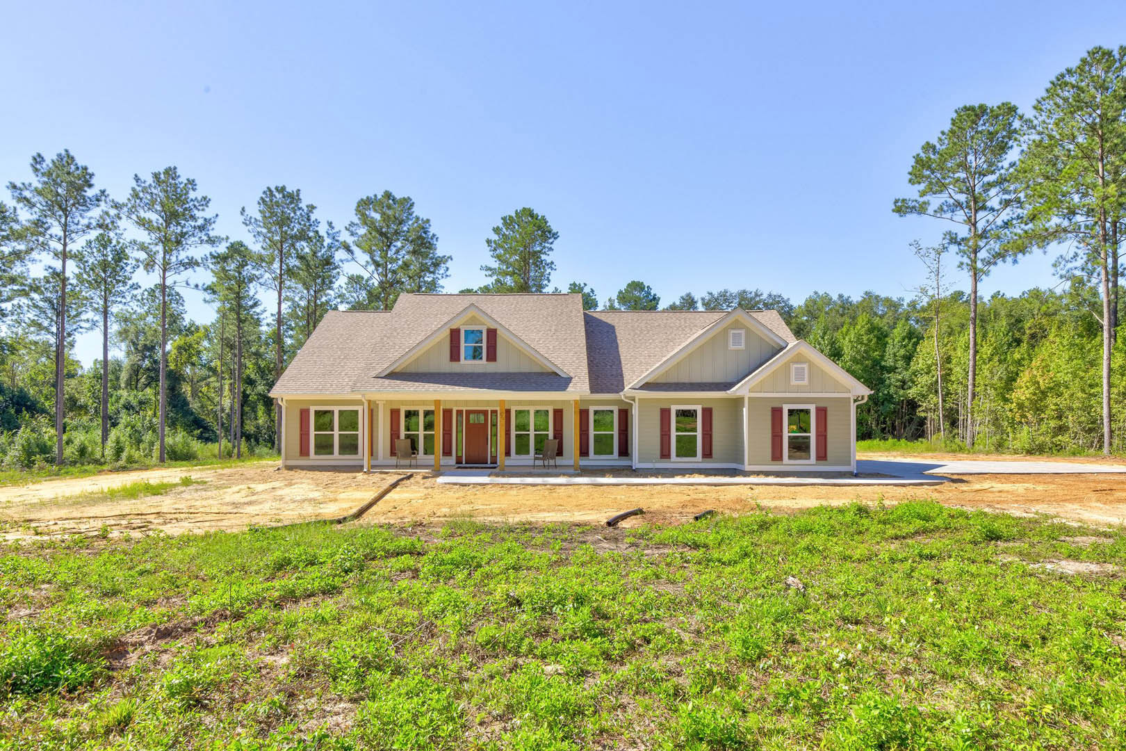 White farmhouse with covered front porch, wooden chair, expansive green lawn, tall leafy trees and dense forest in background, blue sky overhead