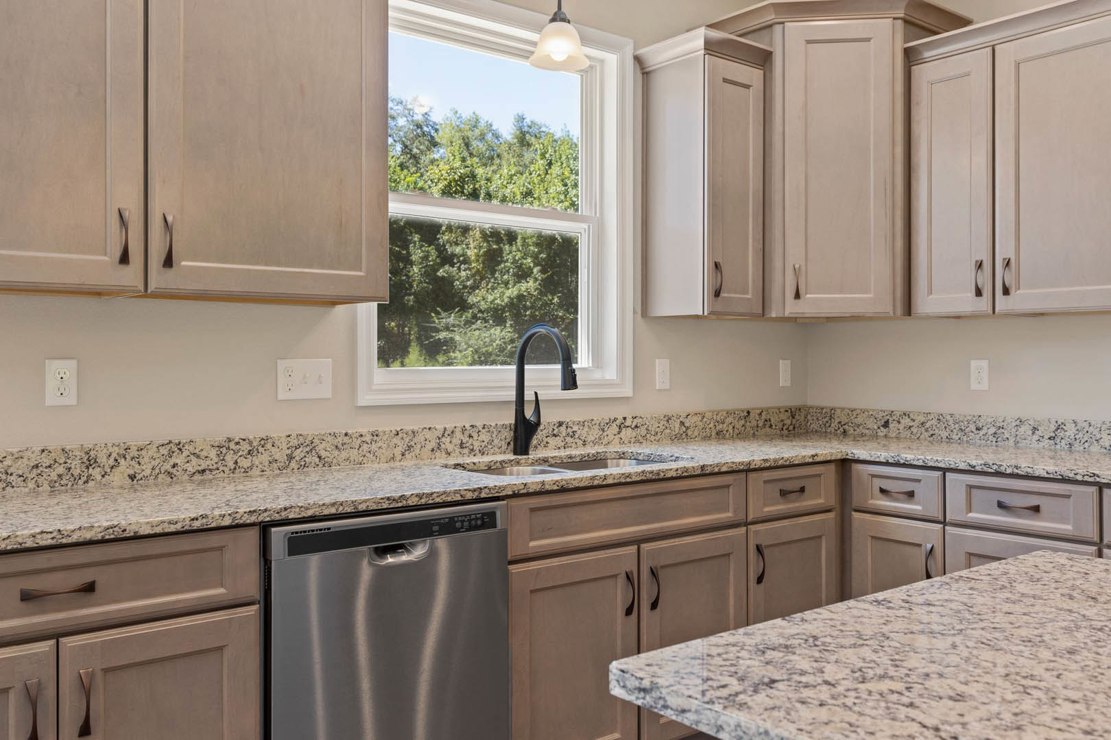 Granite kitchen countertops with undermount sink, black curved faucet, stainless steel dishwasher, white cabinetry, electrical outlet, window overlooking trees