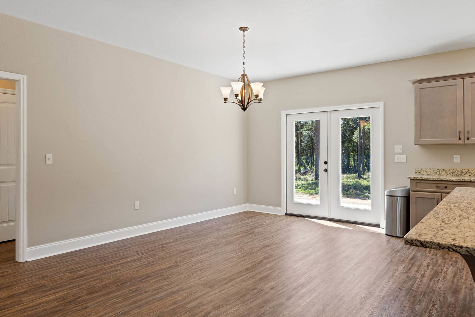 Wood flooring in a spacious room with a glass-paneled double door, ornate chandelier, white cabinetry, and silver decorative container.