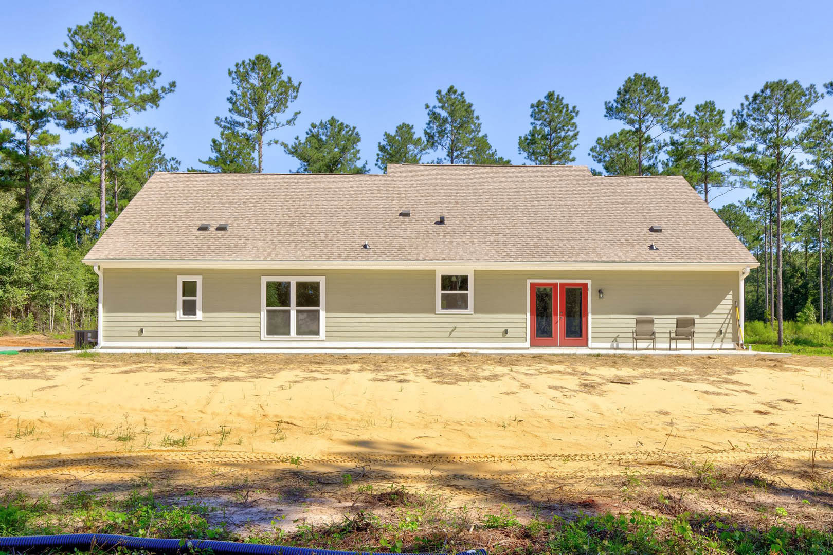 Single-story house with white-framed windows, red double front door with glass panels, tire tracks on sandy dirt road leading to entrance, surrounded by trees and greenery in the