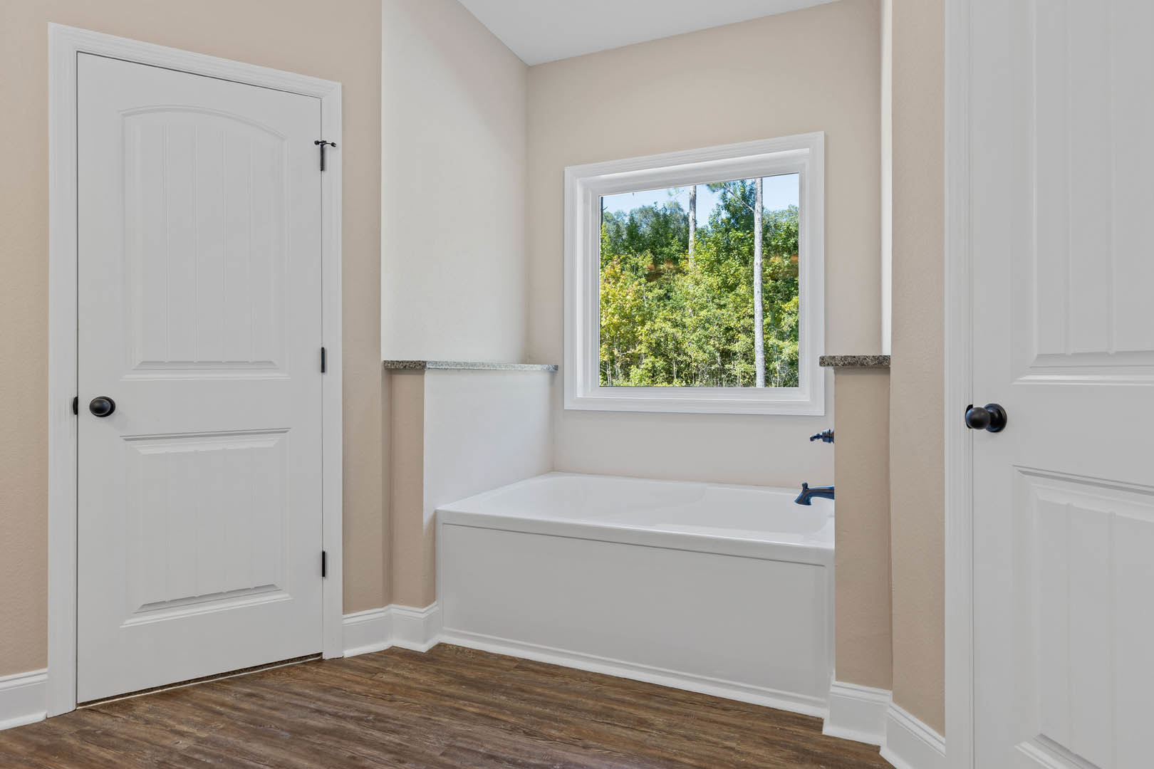 Freestanding white bathtub beneath large window with tree views, wood flooring with white trim, white door featuring black knob