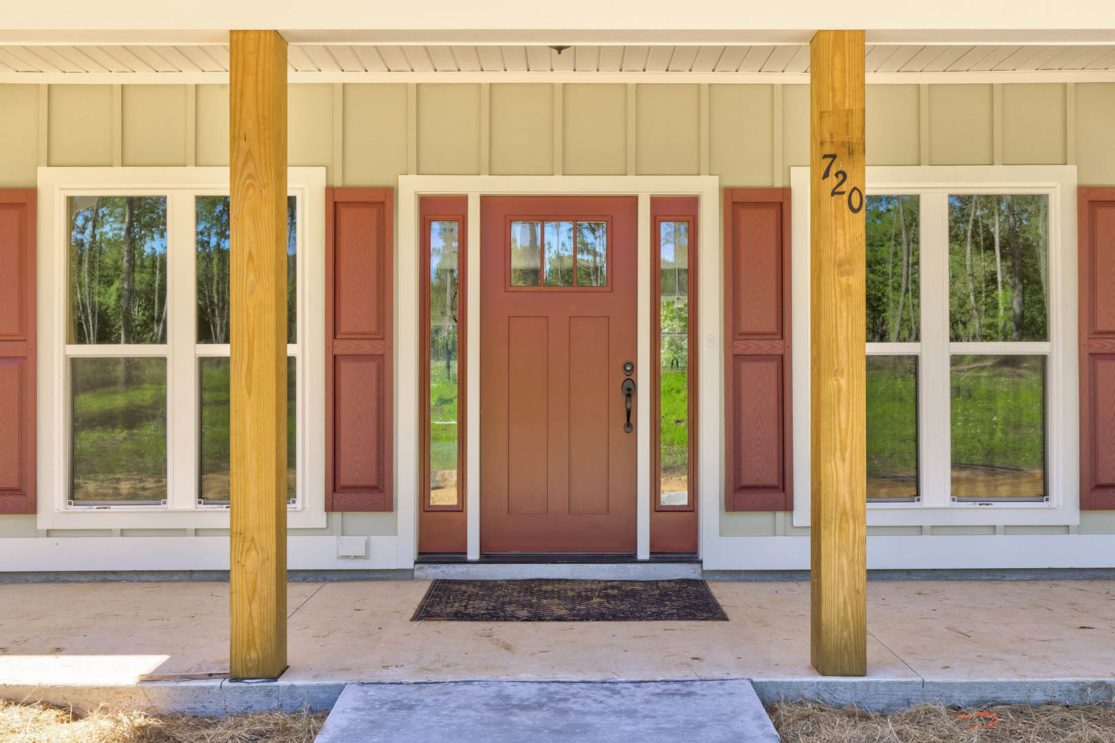 Brown wooden front door with glass panels, brick wall exterior, window reflecting nearby tree, blue sky above, wooden post detail, landscaped entry.