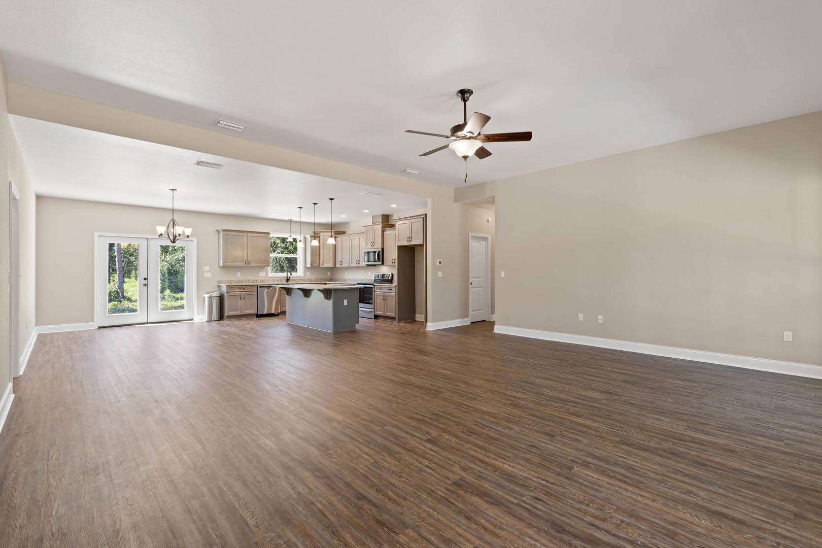 Spacious open floor plan featuring wood flooring, kitchen island, dining area, double doors opening to forest view, ceiling fan with light fixture, and large window overlooking