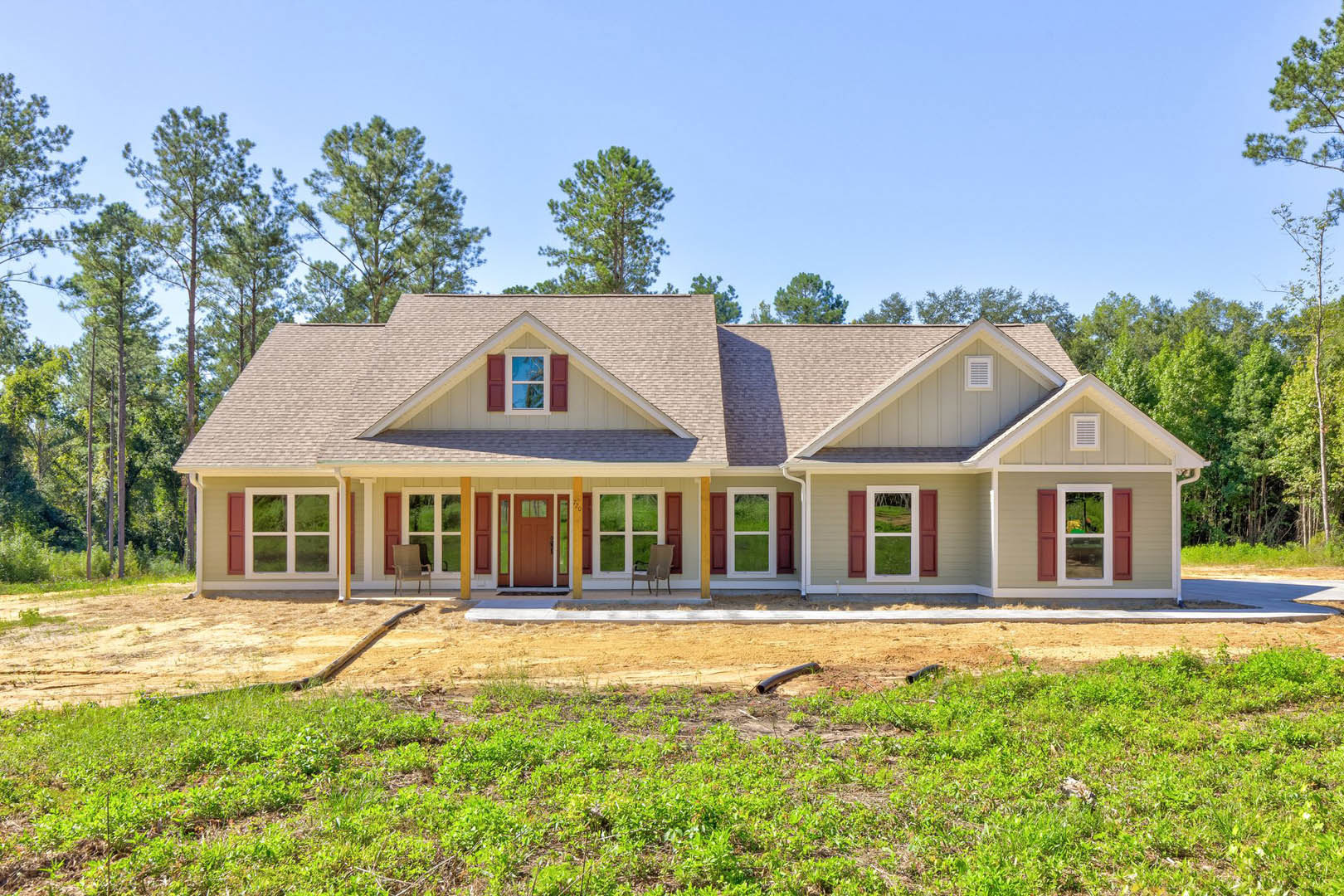 Partially built house with exposed framing, front porch featuring a single chair, grassy yard with visible pipe, door close-up showing unfinished trim, window with red shutters