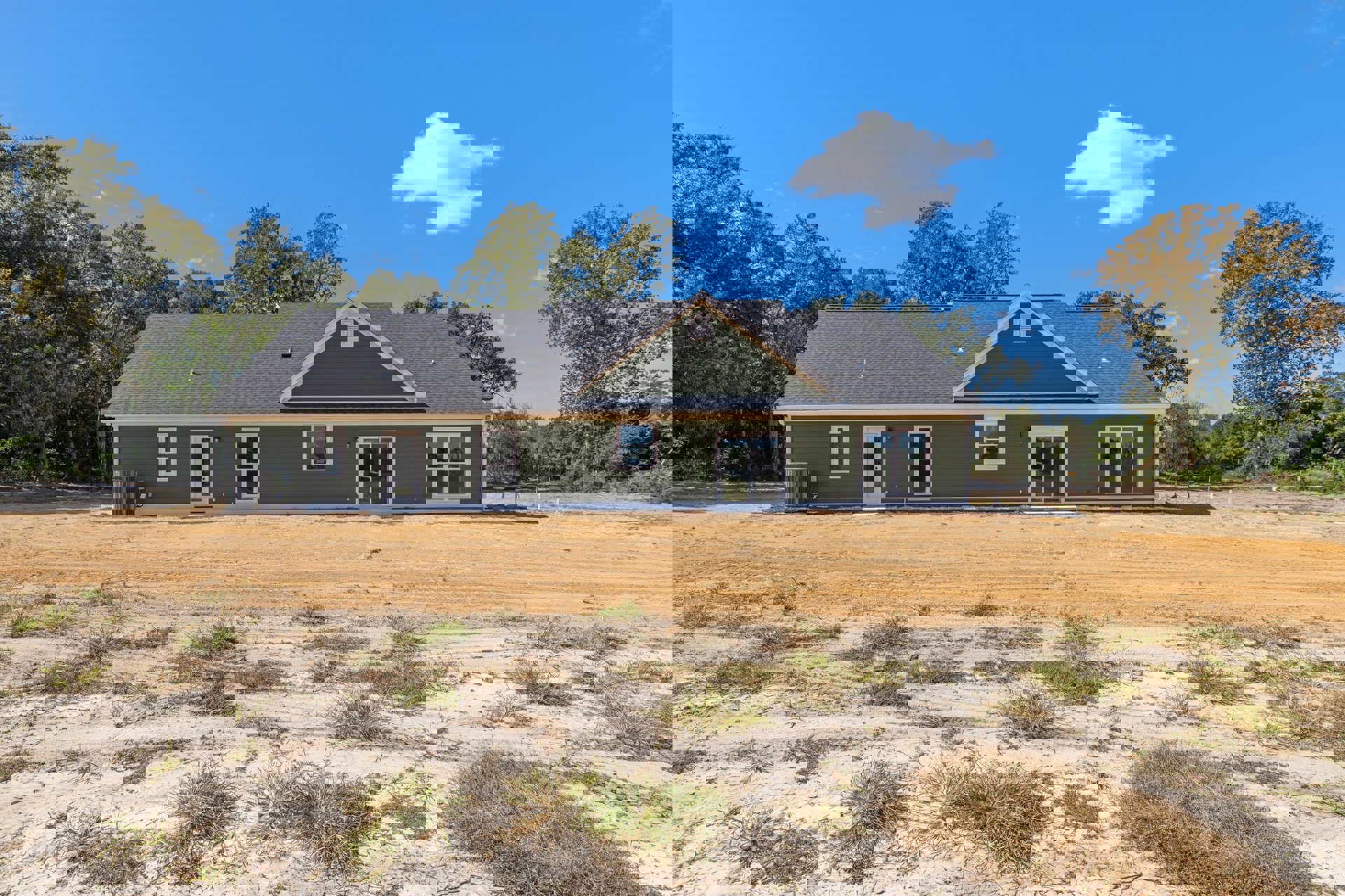 Partially built house with exposed framing, white-trimmed window, sliding glass door, dirt and grass field in foreground, trees and white clouds in blue sky