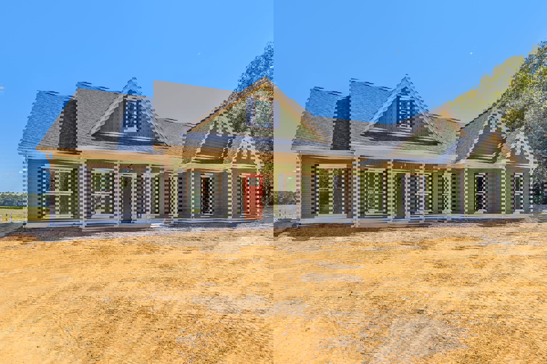 Green house with white pillars, red door with glass pane, white-framed window, dirt field in foreground, blue sky overhead