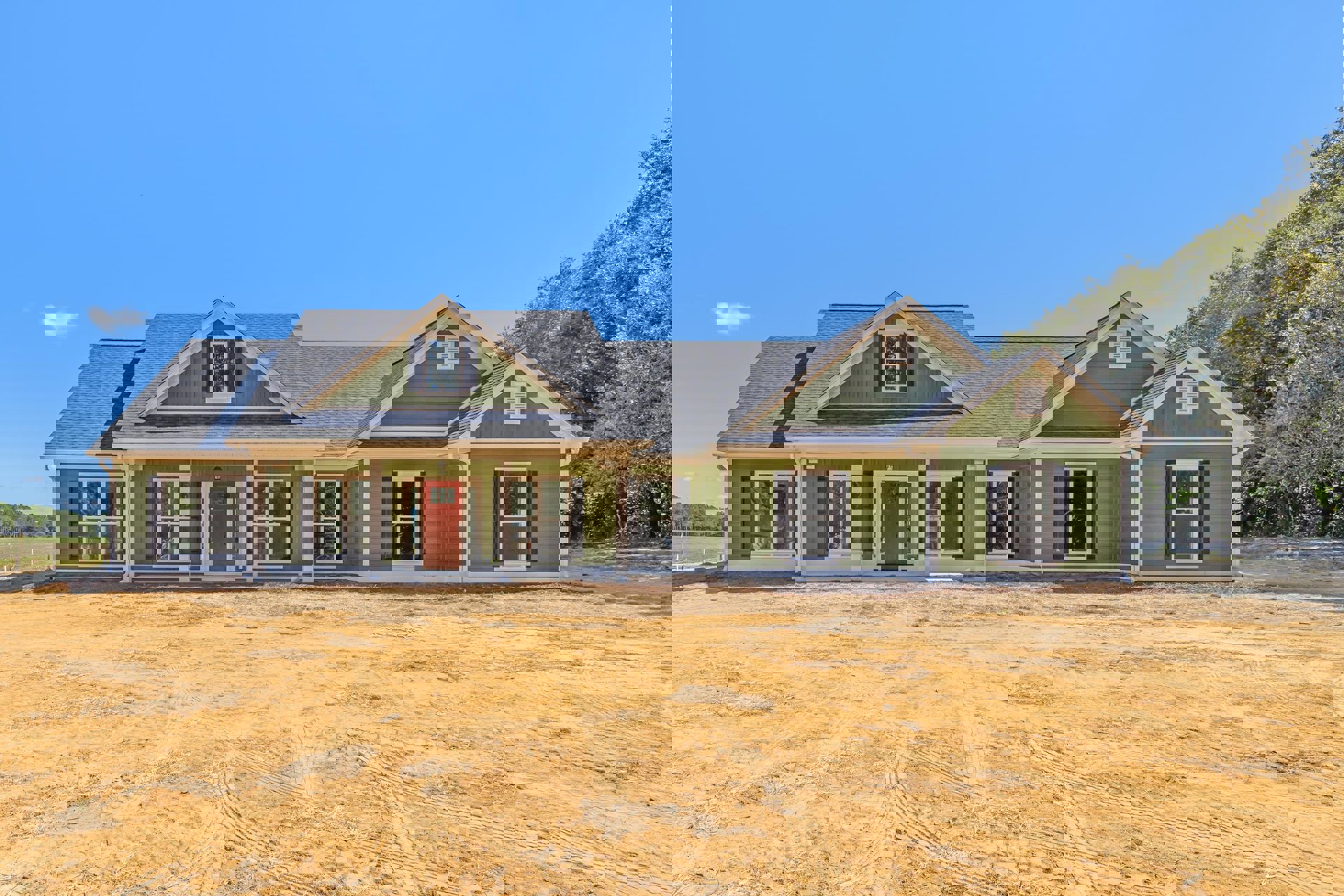 Green house with white trim and red door, white-framed windows including one with broken glass, dirt field with tire tracks in foreground, trees and sky in background