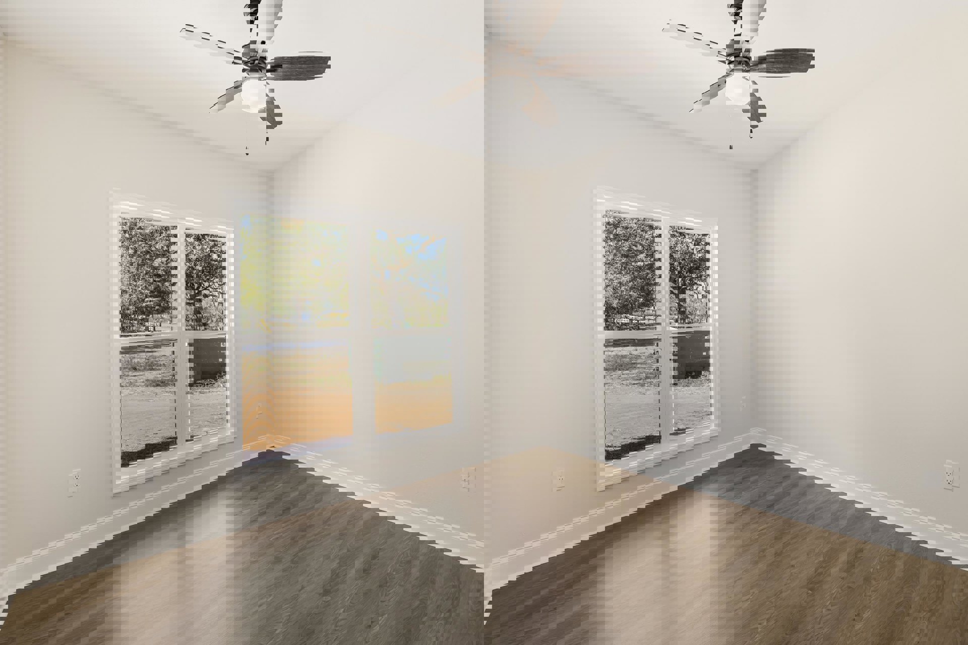 Ceiling fan with light fixture mounted on white plaster ceiling, wood laminate flooring, single window overlooking dirt road and large tree in grassy field.