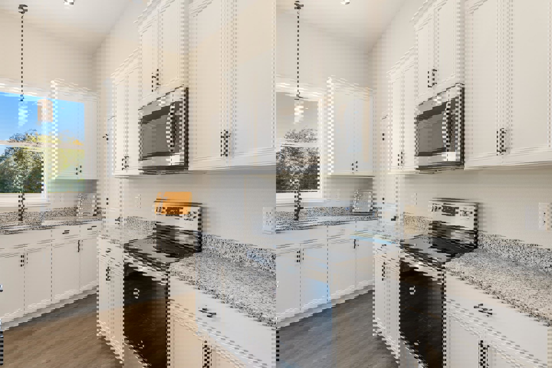 White shaker cabinets, stainless steel microwave and oven, light gray countertops, window with overhead light, wooden gift box with bow on counter