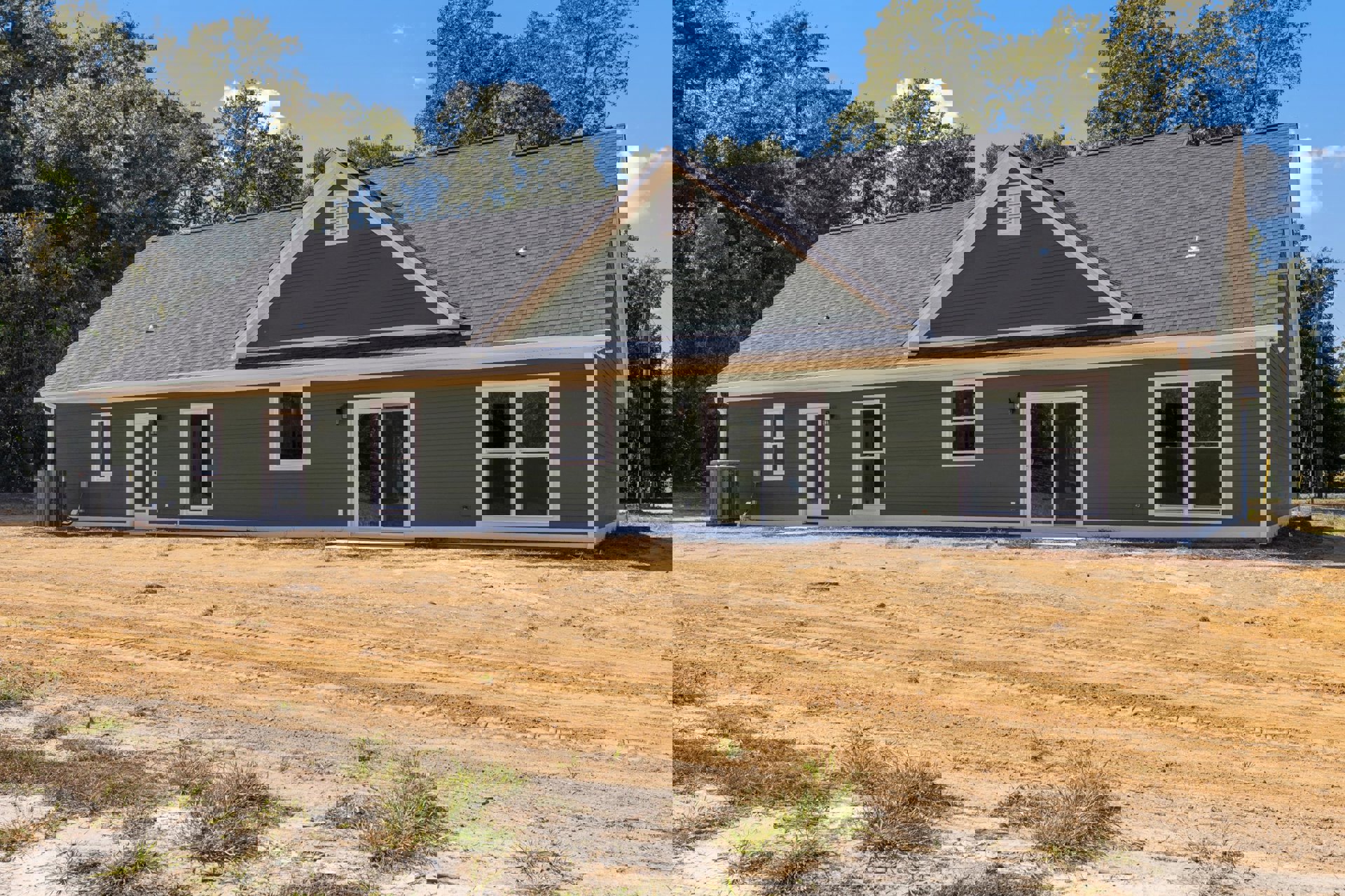 Single-story house with green siding, white door, white-framed window, sliding glass door, and dirt road bordered by grass and trees