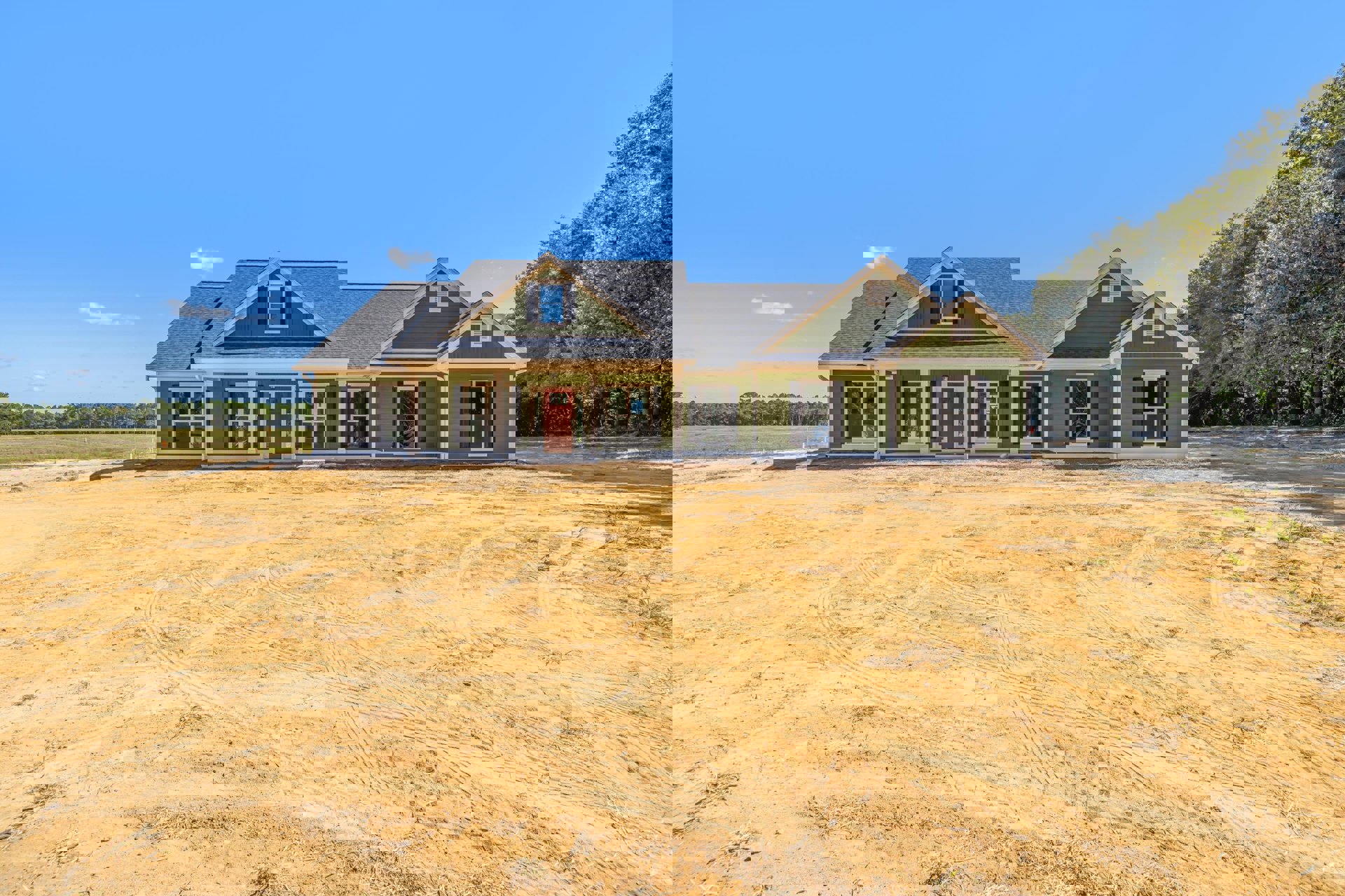 Green house with white-framed windows and red door, surrounded by a large dirt yard with visible tire tracks, under clear blue sky