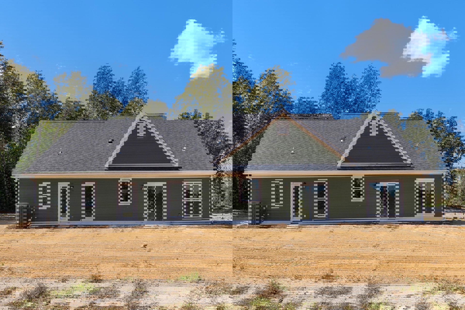 Partially built house with exposed framing, white-framed window, sliding glass door, dirt yard with patches of grass, trees and clouds in the background