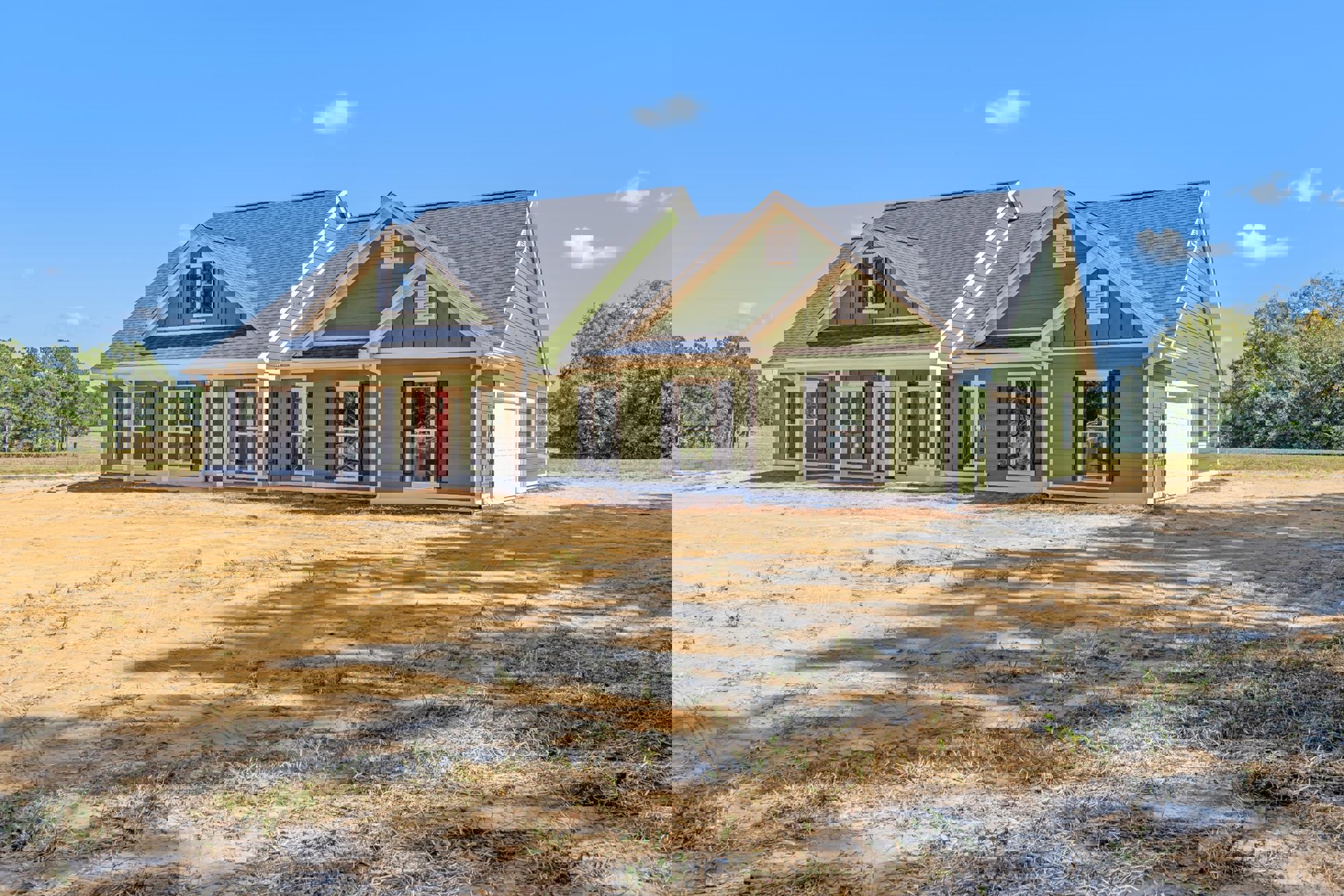 Green custom home with white trim under construction, surrounded by dirt and patches of grass, large windows facing a group of trees, broken glass visible in one window, cloudy sky