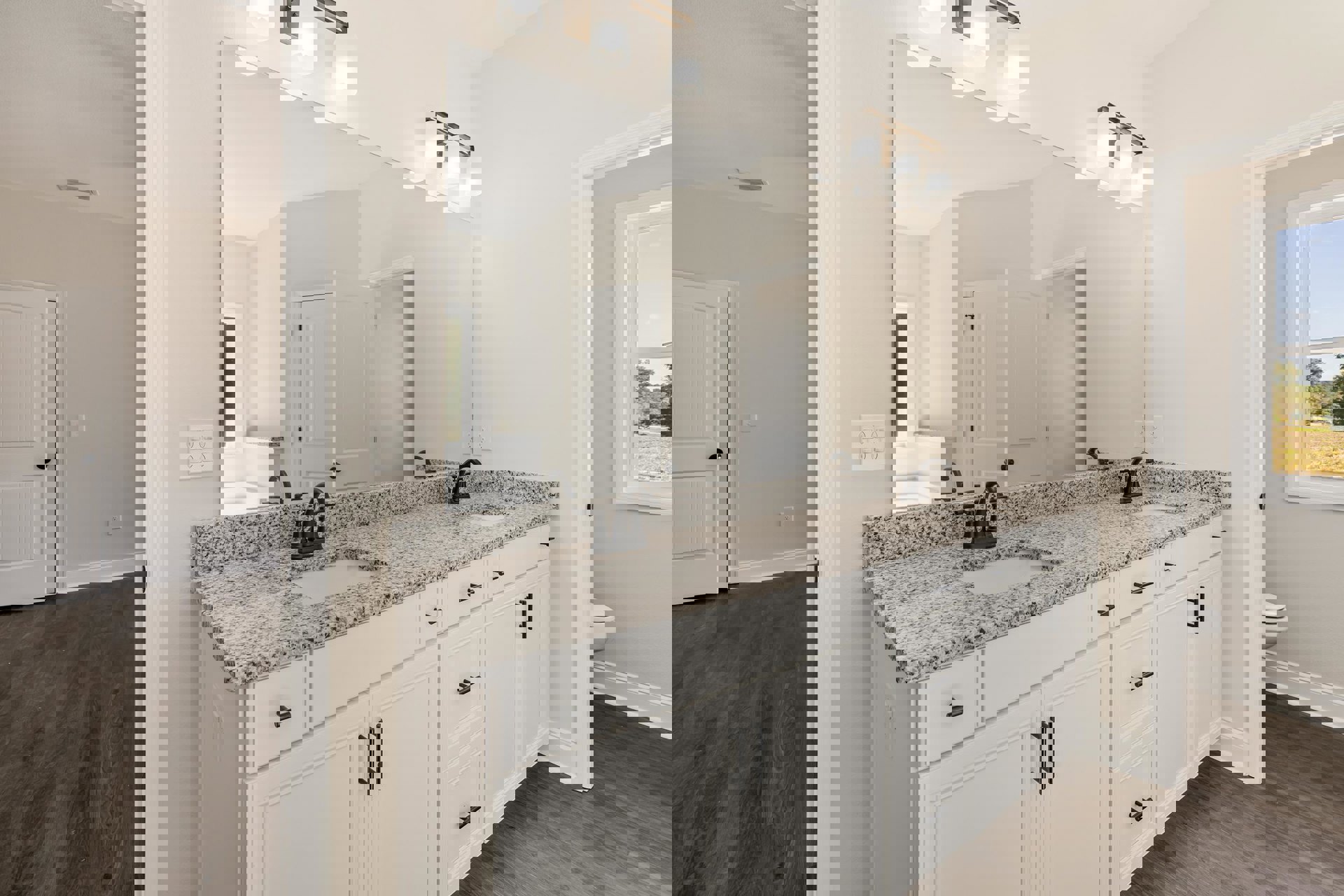 Bathroom with large wall mirror above granite countertop, white cabinets, undermount sink, black faucet, white door with black handle, closed toilet, window showing trees, white