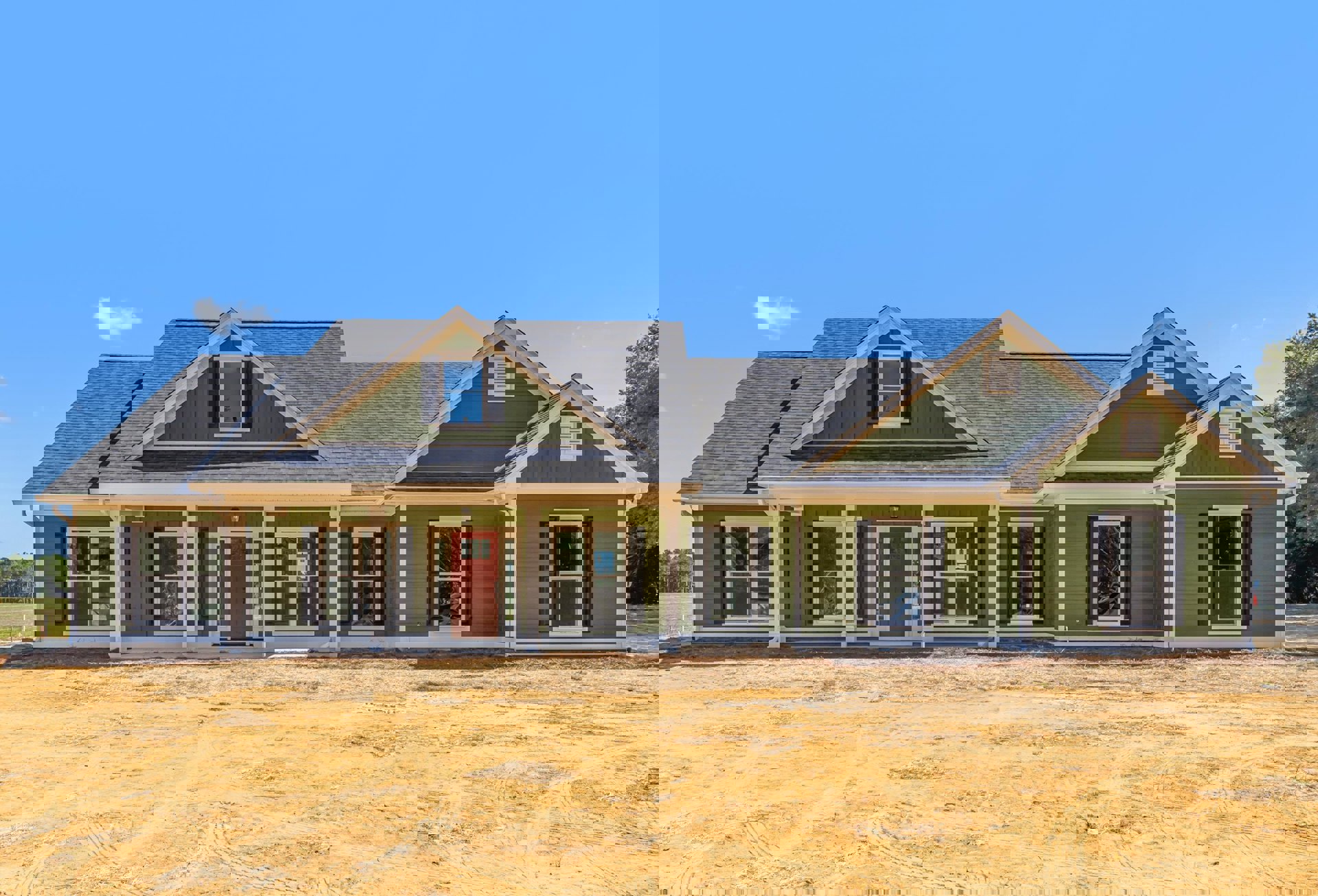 Green siding house with large covered front porch, red door with white trim, multiple windows with white frames, grassy yard bordered by green fence, blue sky overhead.