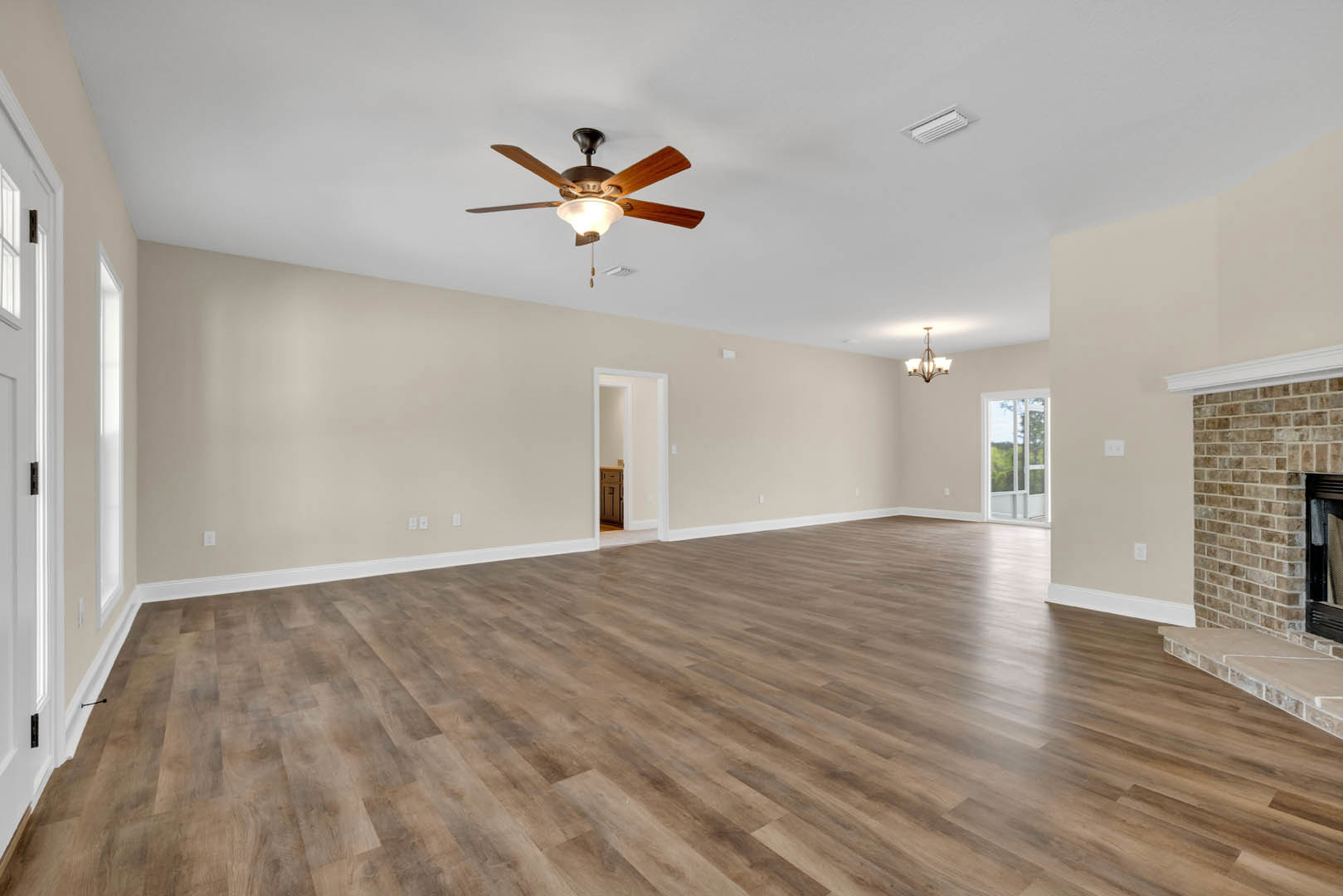 Spacious living area with hardwood flooring, white walls, ceiling fan with light fixture, fireplace, large window overlooking trees, and white door