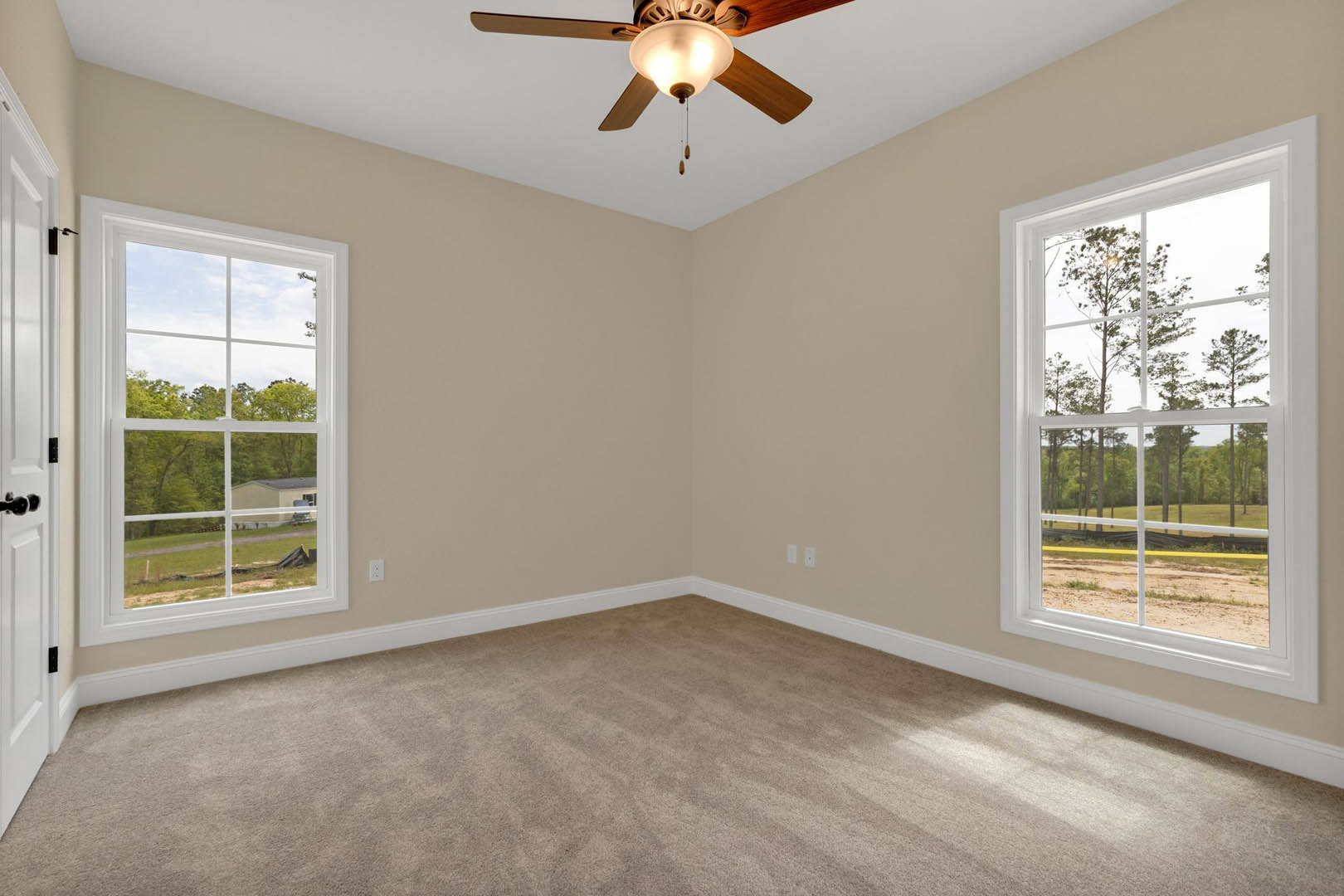 Carpeted room with white walls, ceiling fan with light fixture, large windows showing trees and neighboring house, crown molding along ceiling.