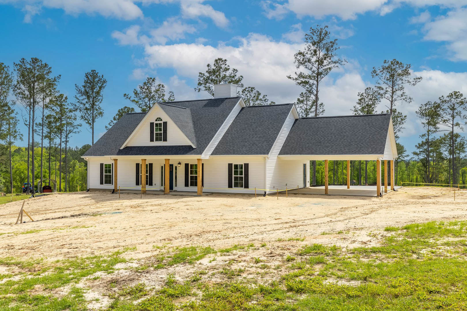 White two-story house with black shutters, large concrete driveway, grassy yard bordered by a yellow rope, mature trees in the background, blue tractor parked near the side