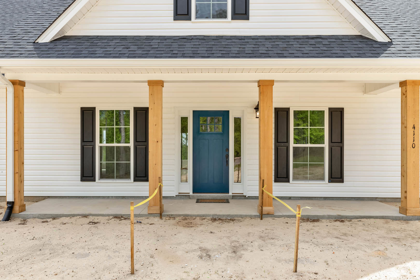 Blue front door with glass window, white-framed windows, gray siding, dirt ground with a doormat, porch area, tree visible outside window, close-up of shingled roof.
