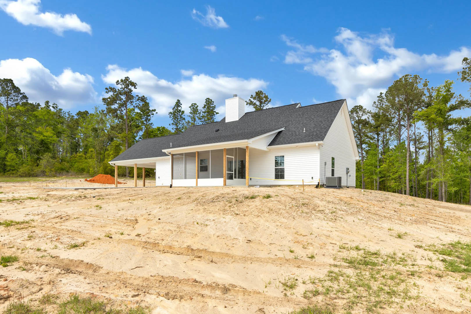 Two-story home with gray siding and white trim, roof with chimney, dirt hill in foreground with tire tracks and red sand pile, yellow caution tape, surrounded by mature trees under