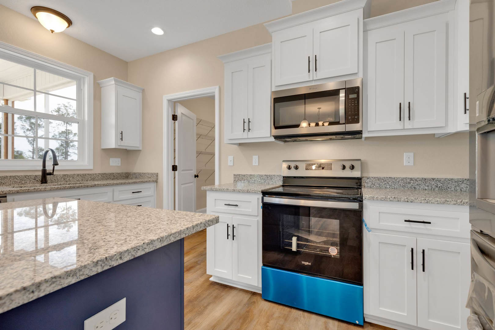 White kitchen cabinets, blue and black oven with glass door, granite countertop, microwave above oven, window with white frame, ceiling light fixture
