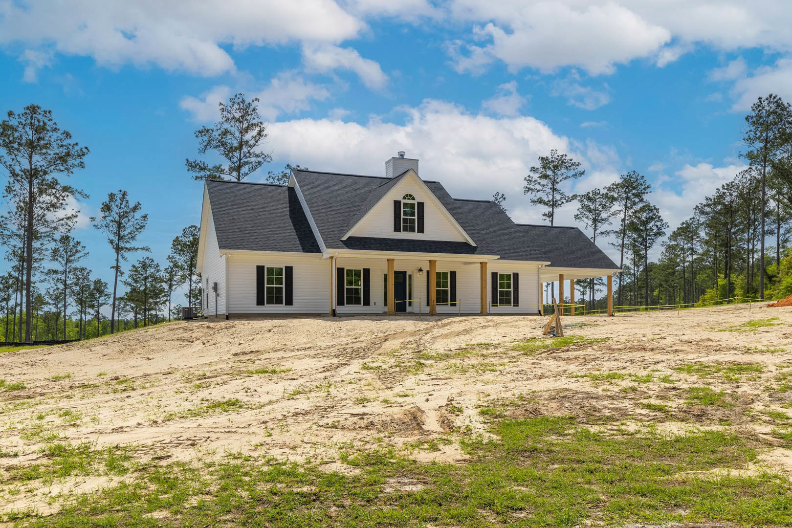 White house with black shutters and black roof, surrounded by a dirt yard and mature trees, cloudy sky overhead