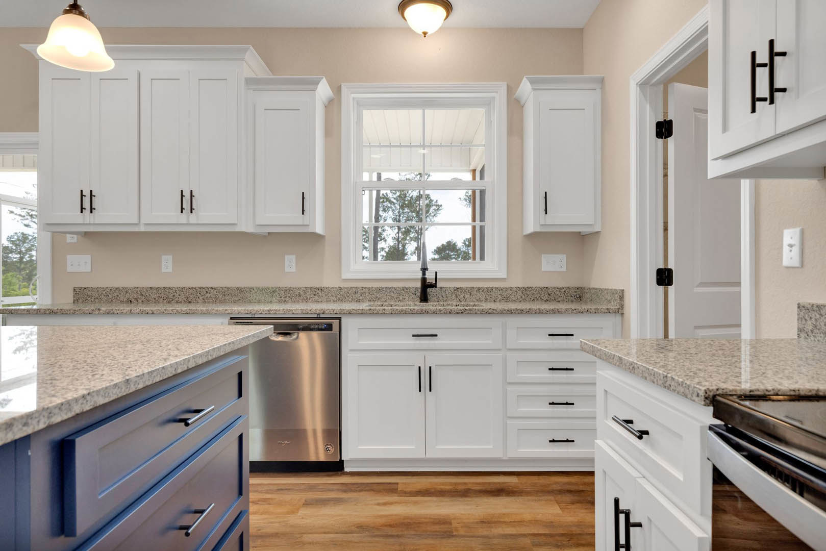 White kitchen cabinets and granite countertops, stainless steel dishwasher beneath counter, wood flooring, stove close-up, window overlooking trees, modern light fixture with