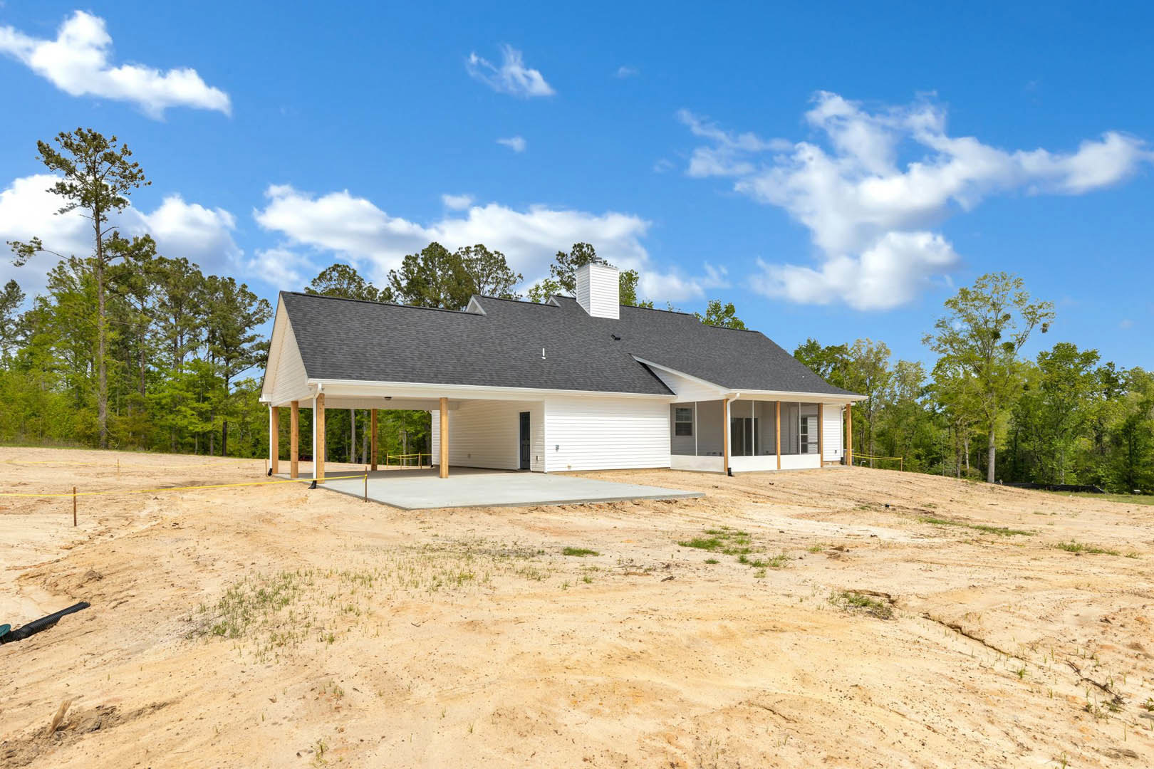 White house with black roof and attached garage, set on a large foundation surrounded by dirt and sparse grass, with green trees and cloudy sky in the background