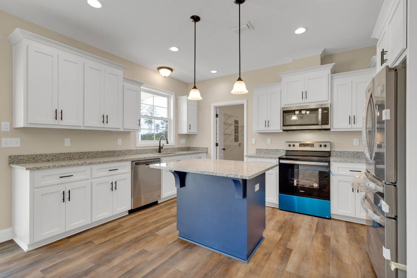 Blue kitchen island with white countertop, stainless steel stove and appliances, row of pendant lights above, white cabinetry, window overlooking trees, hardwood flooring