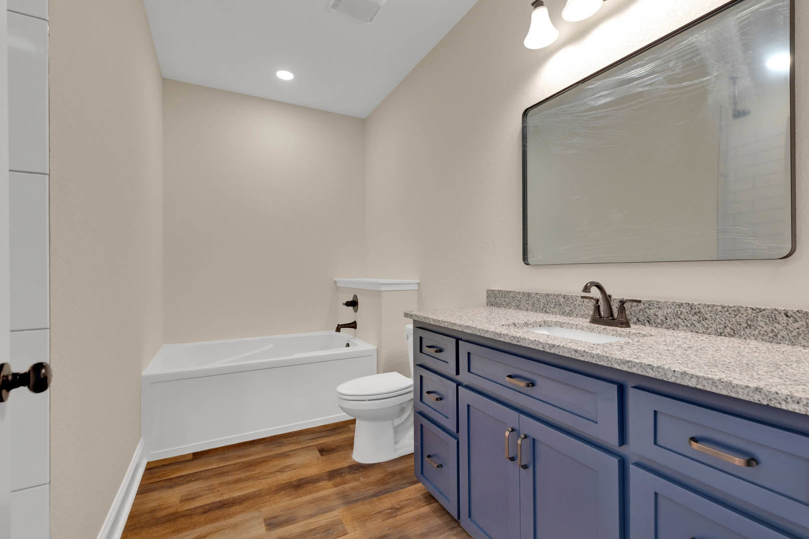 Bathroom with blue vanity featuring long metal handle, white toilet, wood flooring, white walls, white bathtub with wooden base, mirror covered in plastic wrap