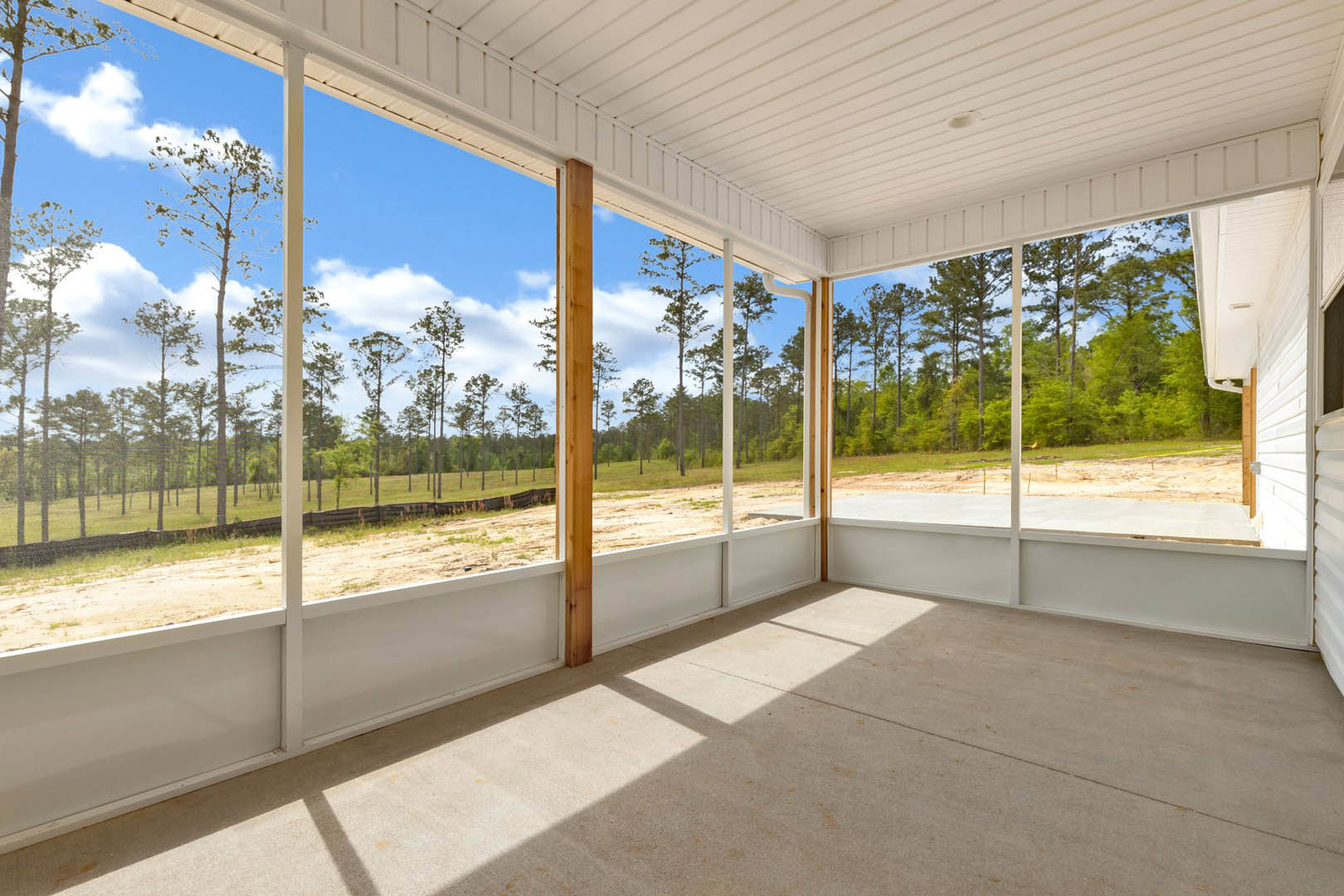 Open living space with expansive windows overlooking lush trees, polished concrete floor, white walls, exposed wood beams, and a prominent wooden post; bright daylight filters