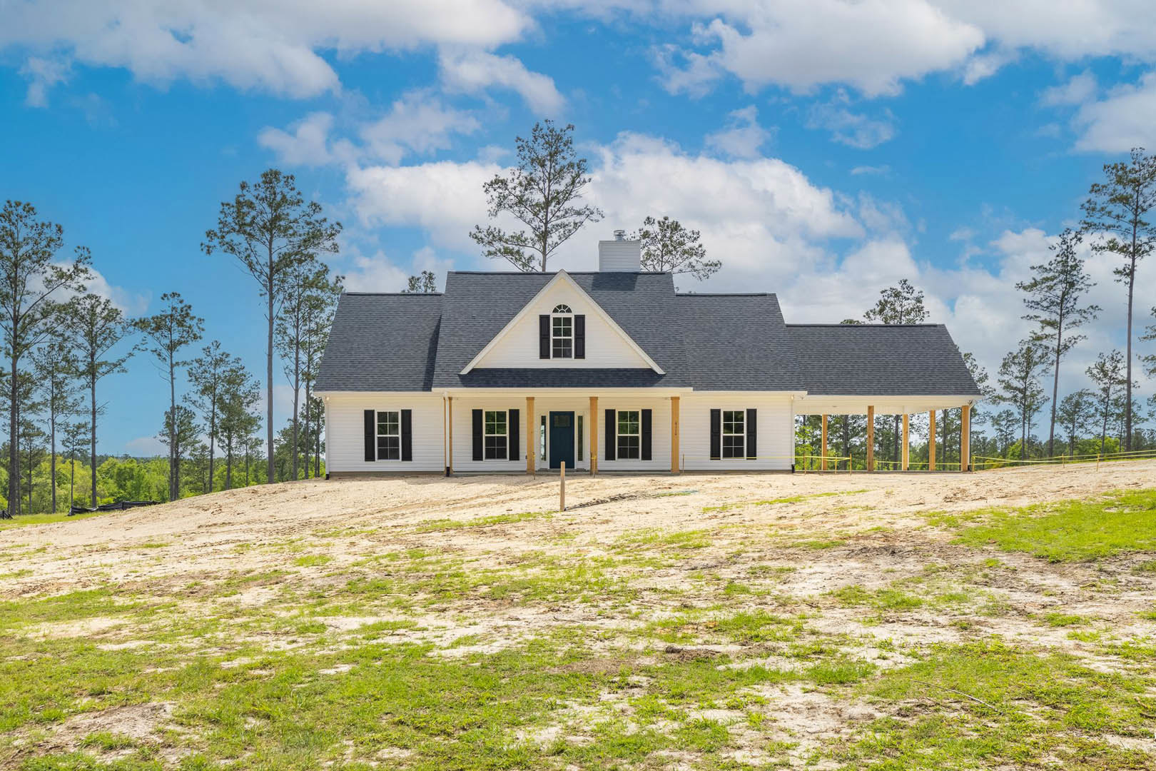 Two-story farmhouse with black roof, blue front door, multi-pane windows, large grassy lawn, and mature leafy tree in front yard under partly cloudy sky
