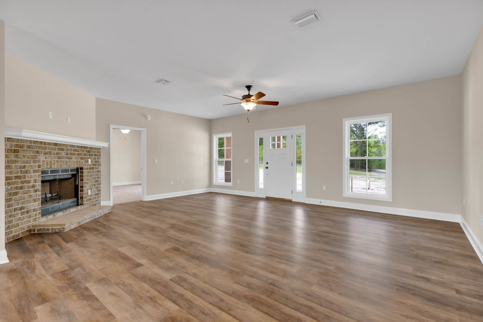 Spacious living room featuring hardwood floors, brick fireplace, ceiling fan with light, large window overlooking trees, and white door with glass panes