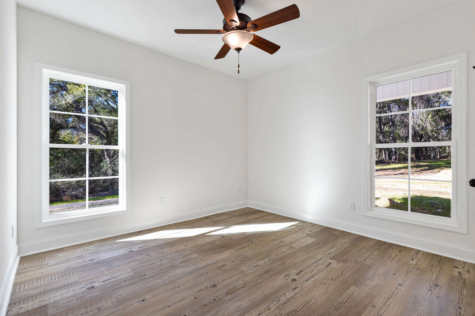 Ceiling fan with light fixture mounted on white ceiling, large windows overlooking trees, wood flooring, white walls