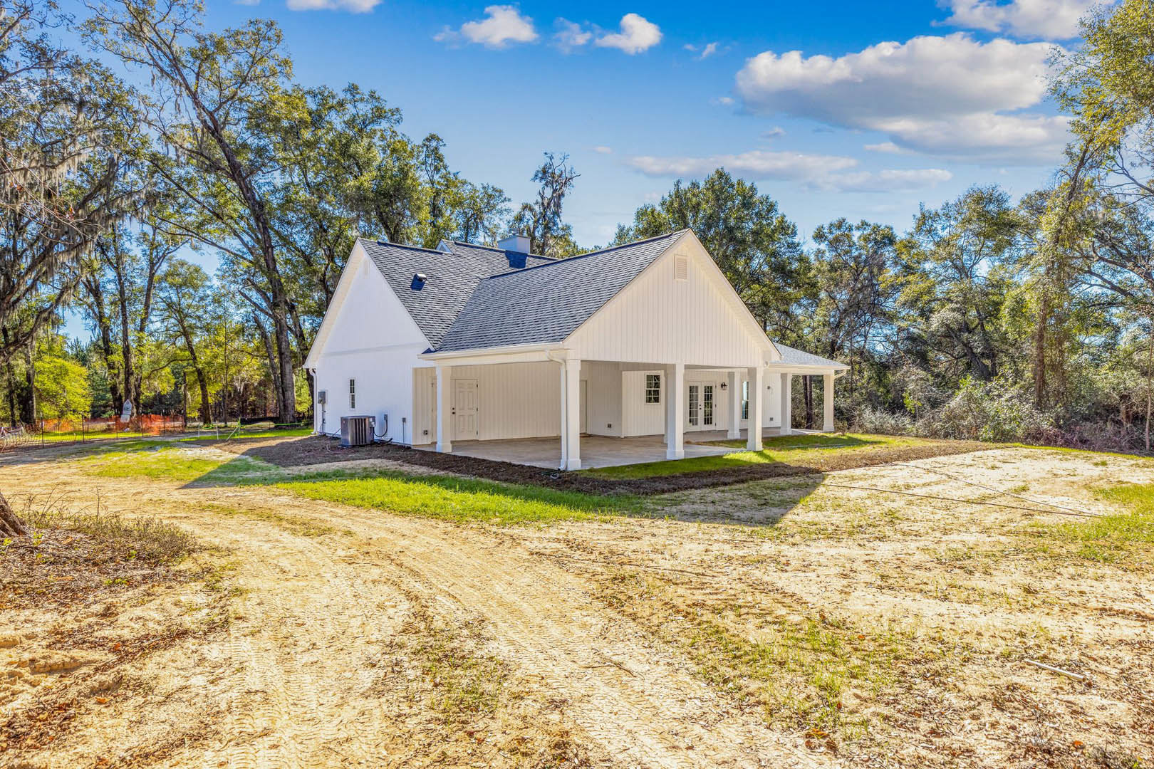 White cottage-style house with covered porch, surrounded by trees, dirt road and grassy yard in foreground, cloudy sky above