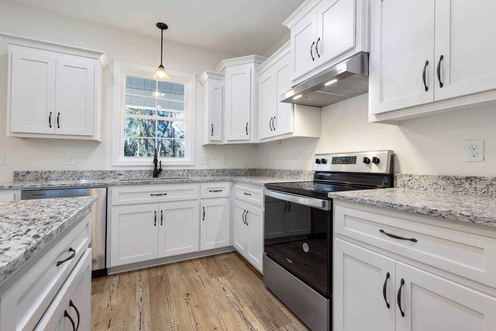 White kitchen cabinets with granite countertops, stainless steel oven, ceiling light above a window, white electrical outlet, close-up of cabinet drawer handle.