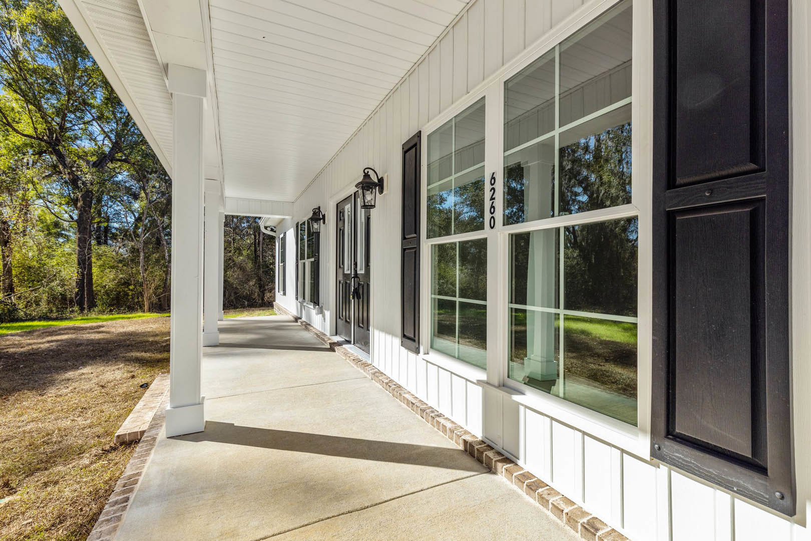 White house exterior with black trim, spacious porch, large front window, green lawn, brick edging, and leafy tree in foreground