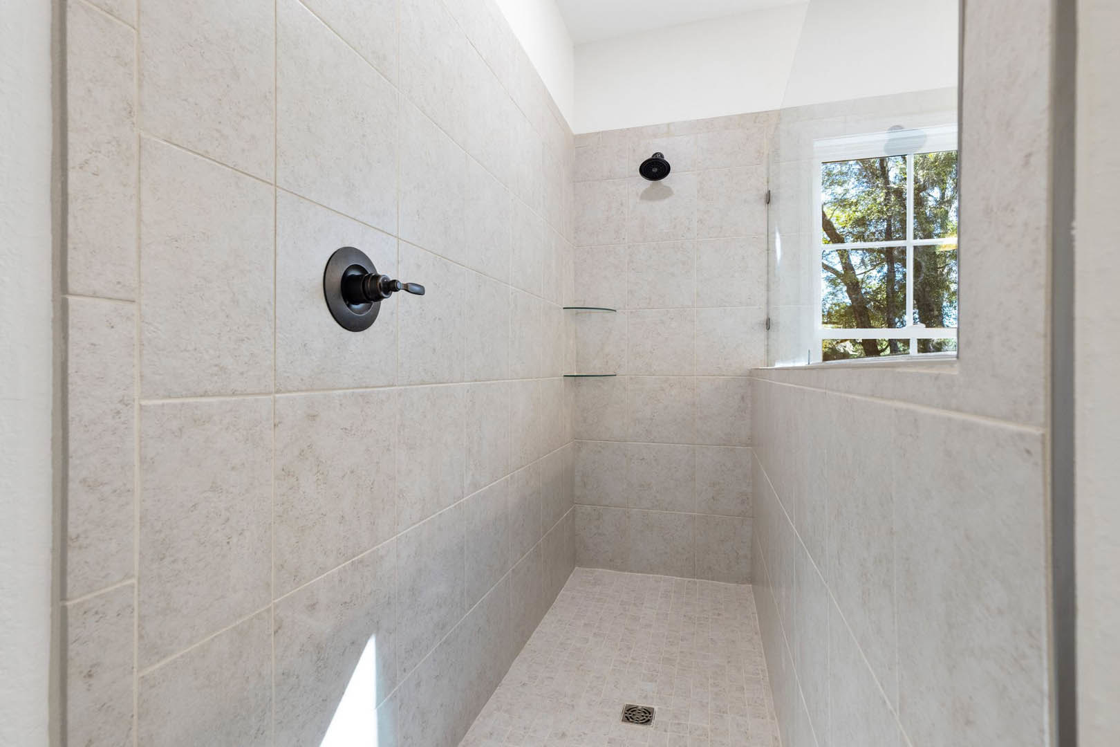 Modern bathroom shower with dual chrome shower heads, white tile walls, glass shelf, window overlooking trees, and silver shower drain