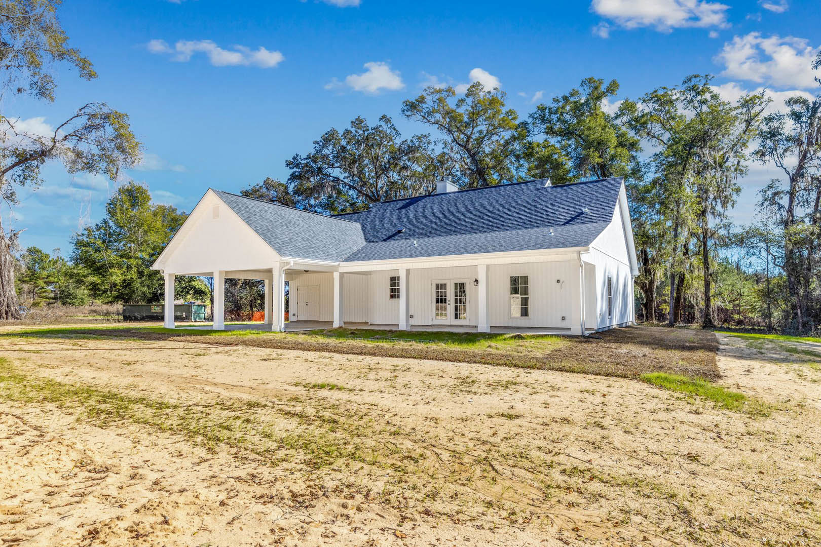 White house with blue roof and expansive front porch, surrounded by green grass and mature trees under a partly cloudy sky