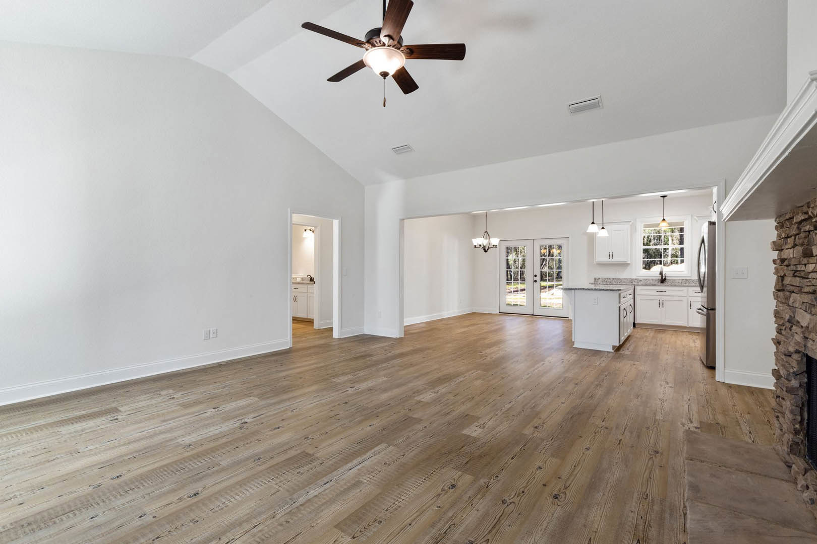 Spacious living area with wood flooring, ceiling fan with light fixture, double glass-paneled doors, and white kitchen island topped with marble countertops