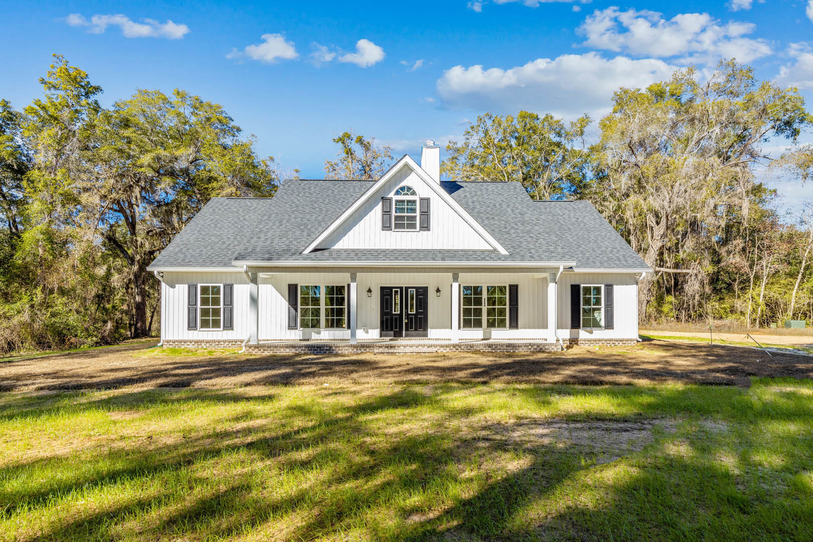 White house with large front porch, black double doors, expansive green lawn, mature trees in background, blue sky with scattered clouds