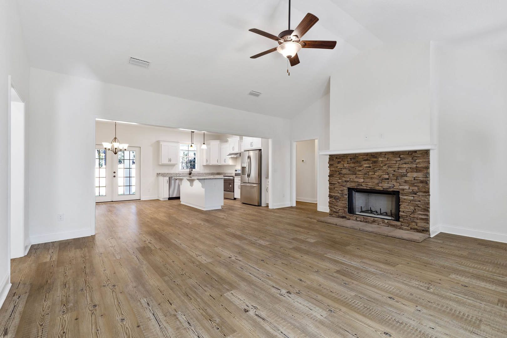 Open-concept living room with wood flooring, stone fireplace with glass window, ceiling fan with light, and adjacent kitchen featuring stainless steel refrigerator