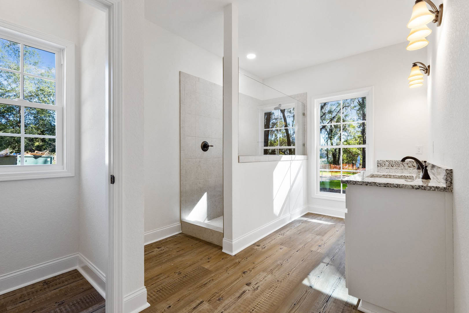 Bathroom with glass shower enclosure, white sink vanity, wood flooring, tile walls, large window overlooking trees, modern lamp fixture