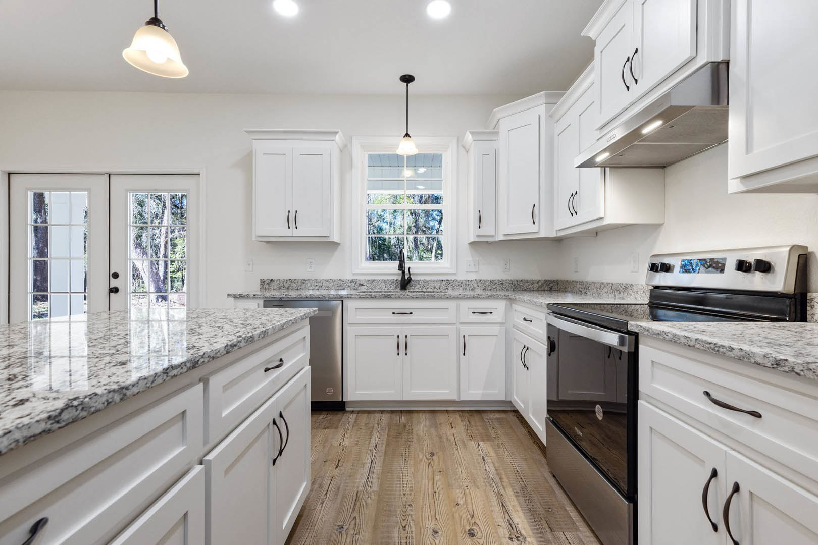 White kitchen with granite countertops, black cabinet handles, stainless steel stove, ceiling light fixture, and window letting in natural light.