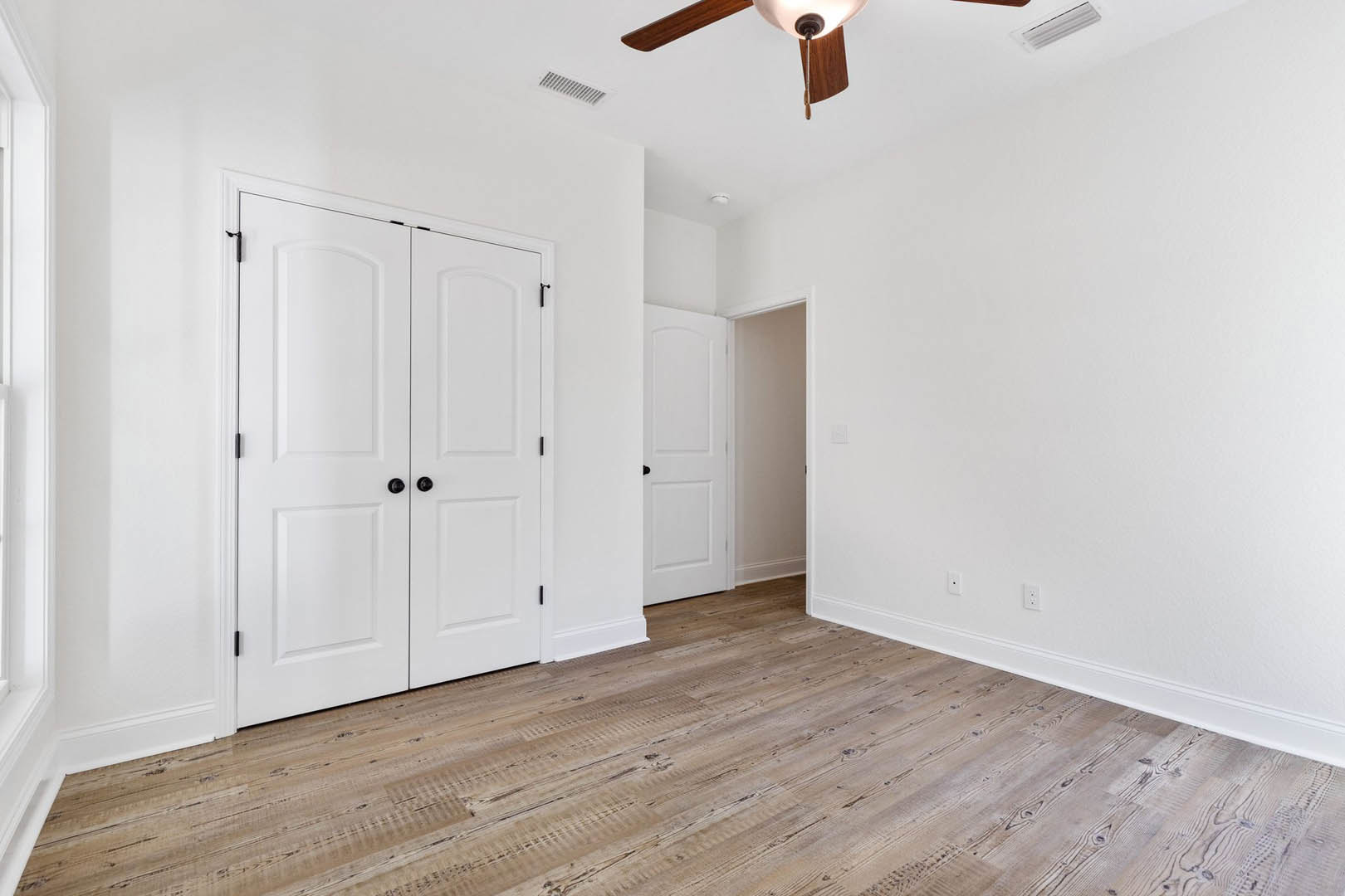 Room with wood flooring, white walls, white closet doors with black knobs, white entry door with black handle, ceiling fan, and close-up of a modern light fixture