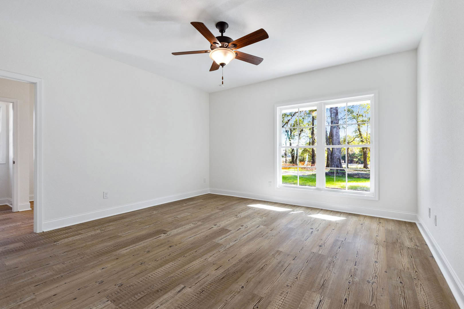 Bedroom with wood flooring, white plaster walls, ceiling fan with integrated light fixture, large window overlooking trees.