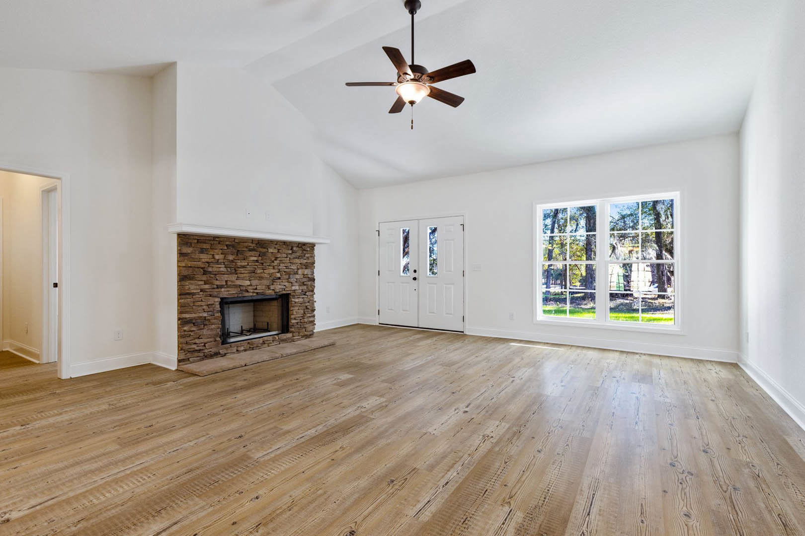 Living room with wood flooring, brick fireplace, ceiling fan with light, double white doors with glass panes, large window showing trees outside