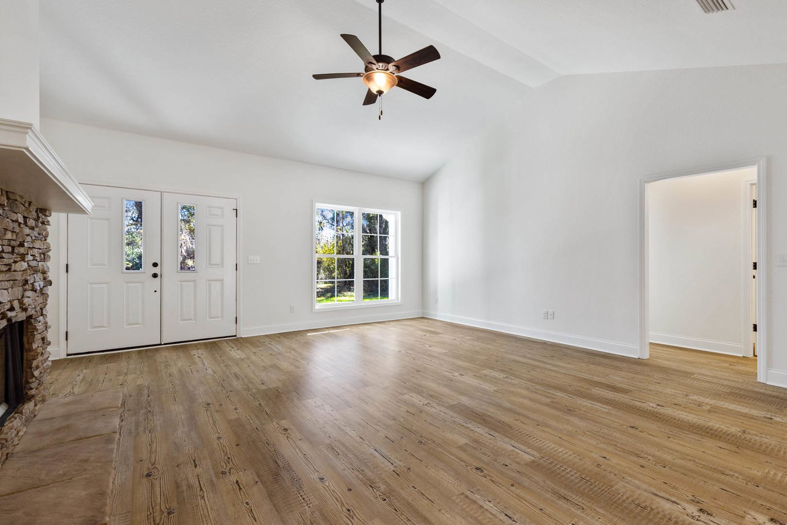Ceiling fan with light fixture, hardwood floor, large window showing trees, white double doors with glass panes, white single door, neutral walls